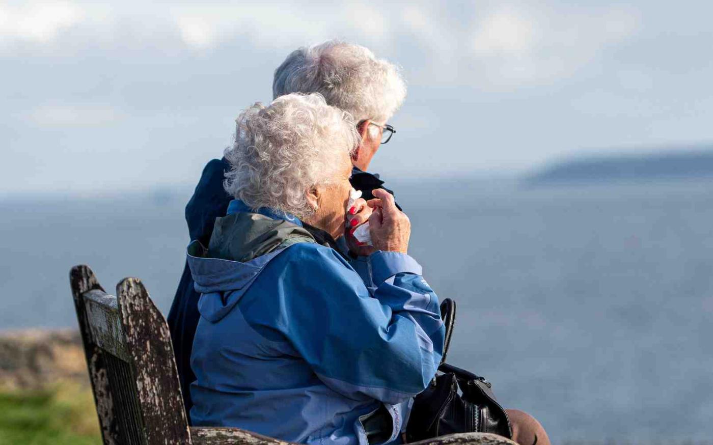 Two elderly people sitting on a bench looking at the sea